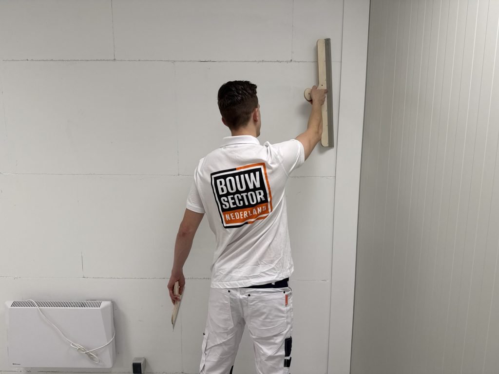 Stukadoor met het logo van Bouwsector Nederland op zijn rug zet een lange spaan hoog tegen een wand