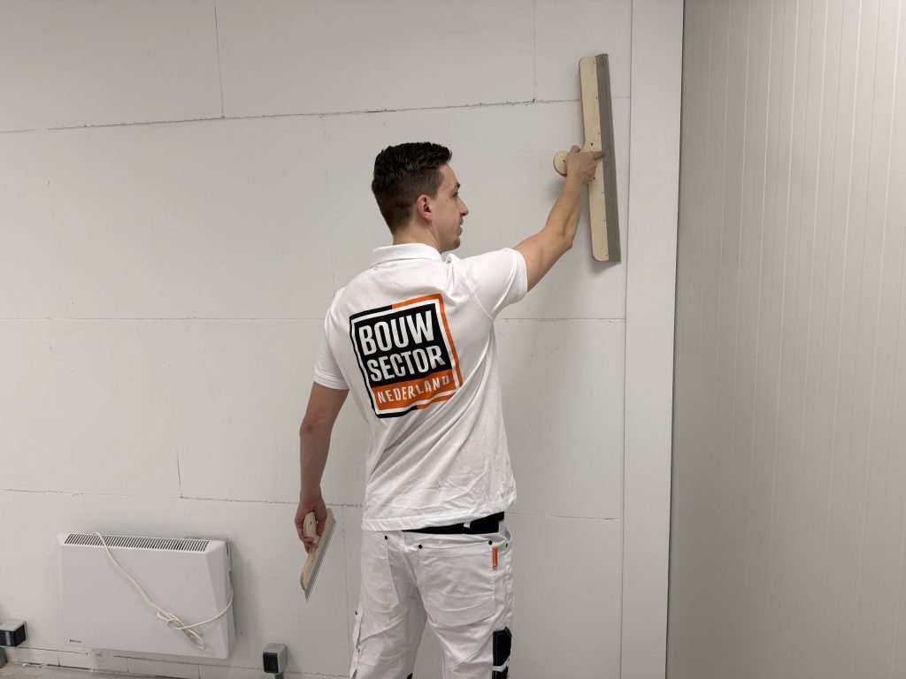 Stukadoor kijkt opzij terwijl hij met een lange spaan een wand vlak zet, plamuurmes in zijn linkerhand